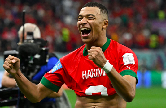 Kylian Mbappe with both hands raised in fists of joy, smiling wide and wearing a half-pulled on red Moroccan jersey that says Hakimi.